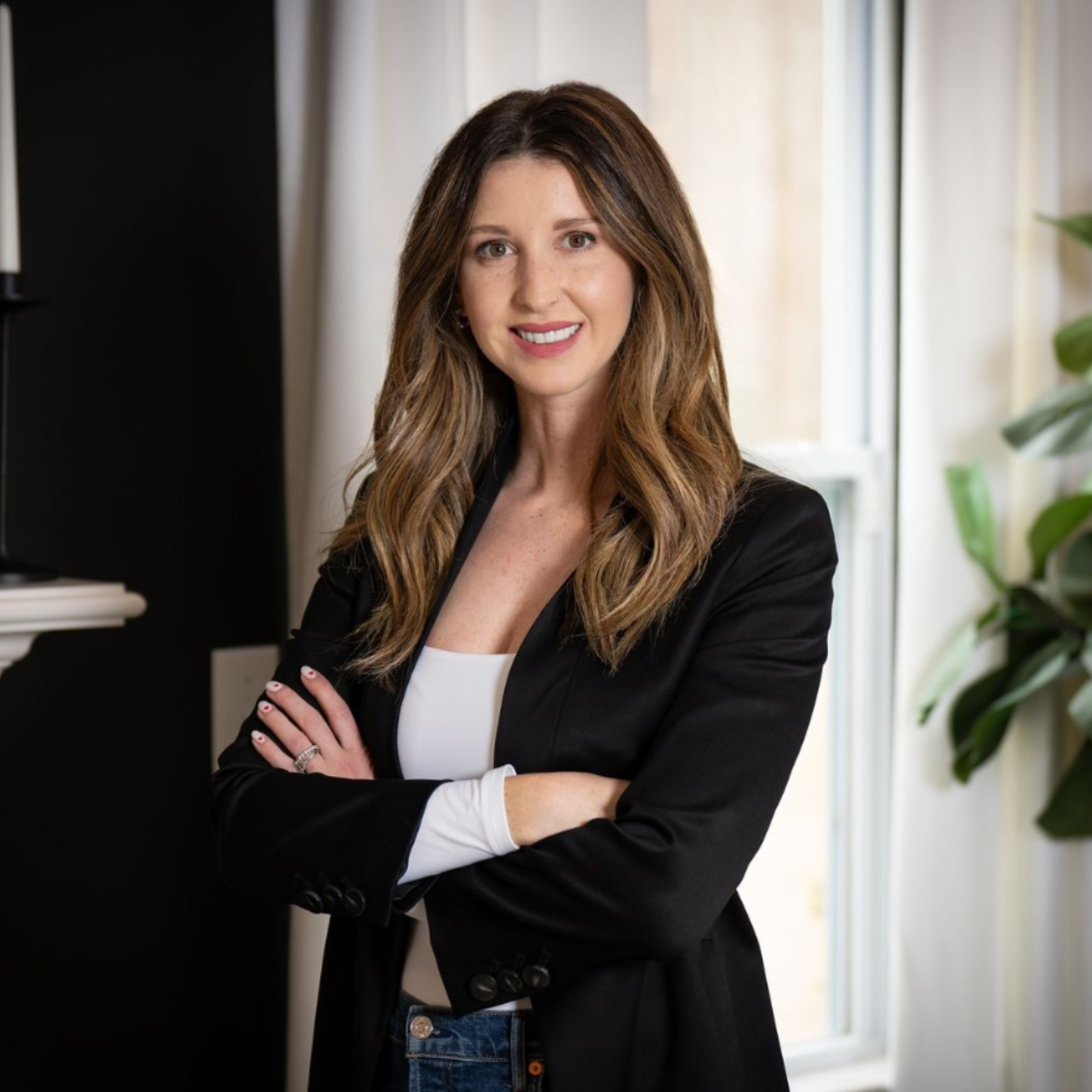 A woman in a black jacket is standing in front of a window with her arms crossed.