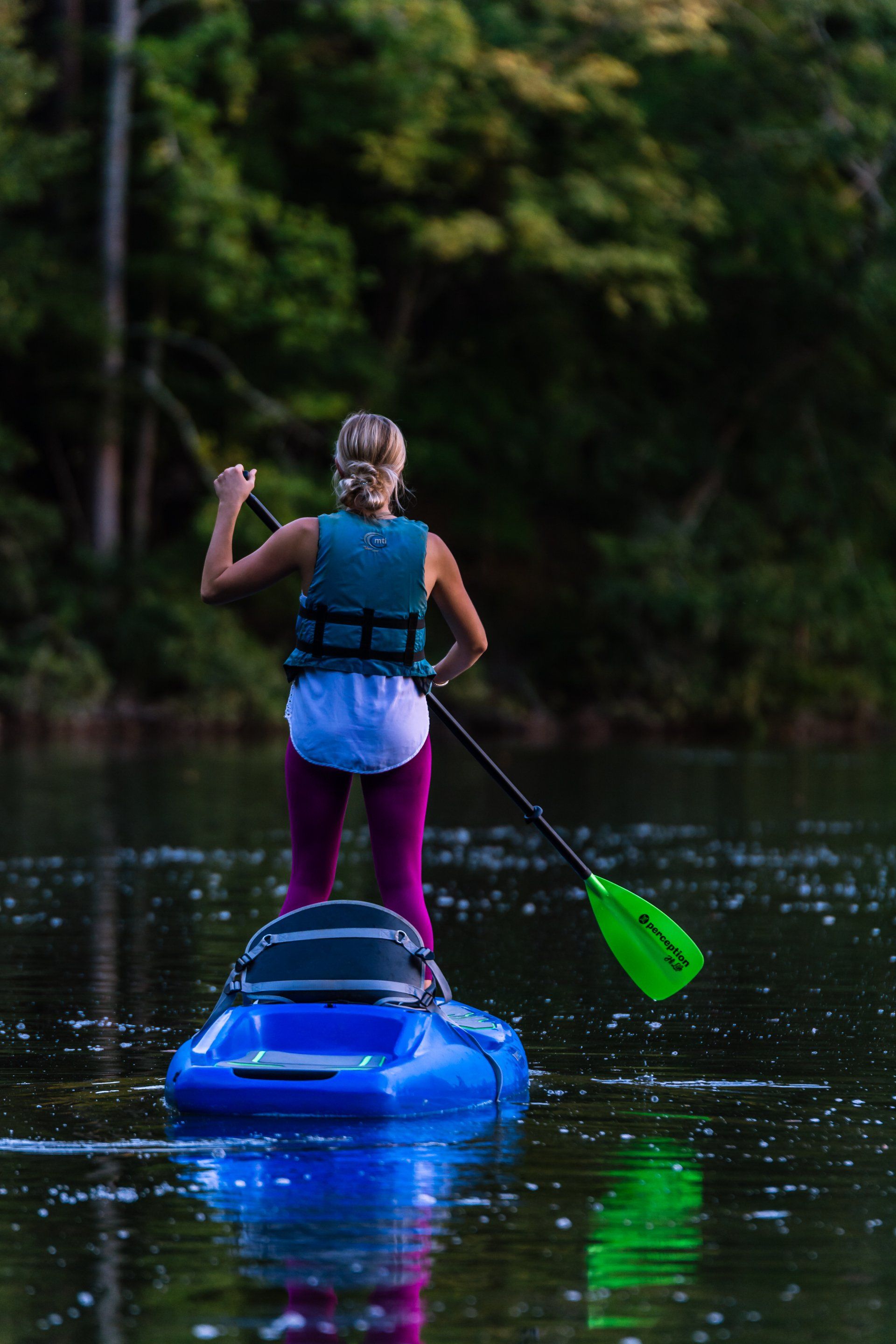 Woman paddleboarding on a blue inflatable board; green paddle, lake at dusk.