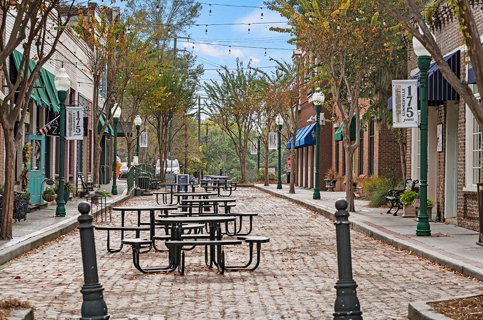 Cobblestone street lined with shops, picnic tables, and trees under a blue sky.
