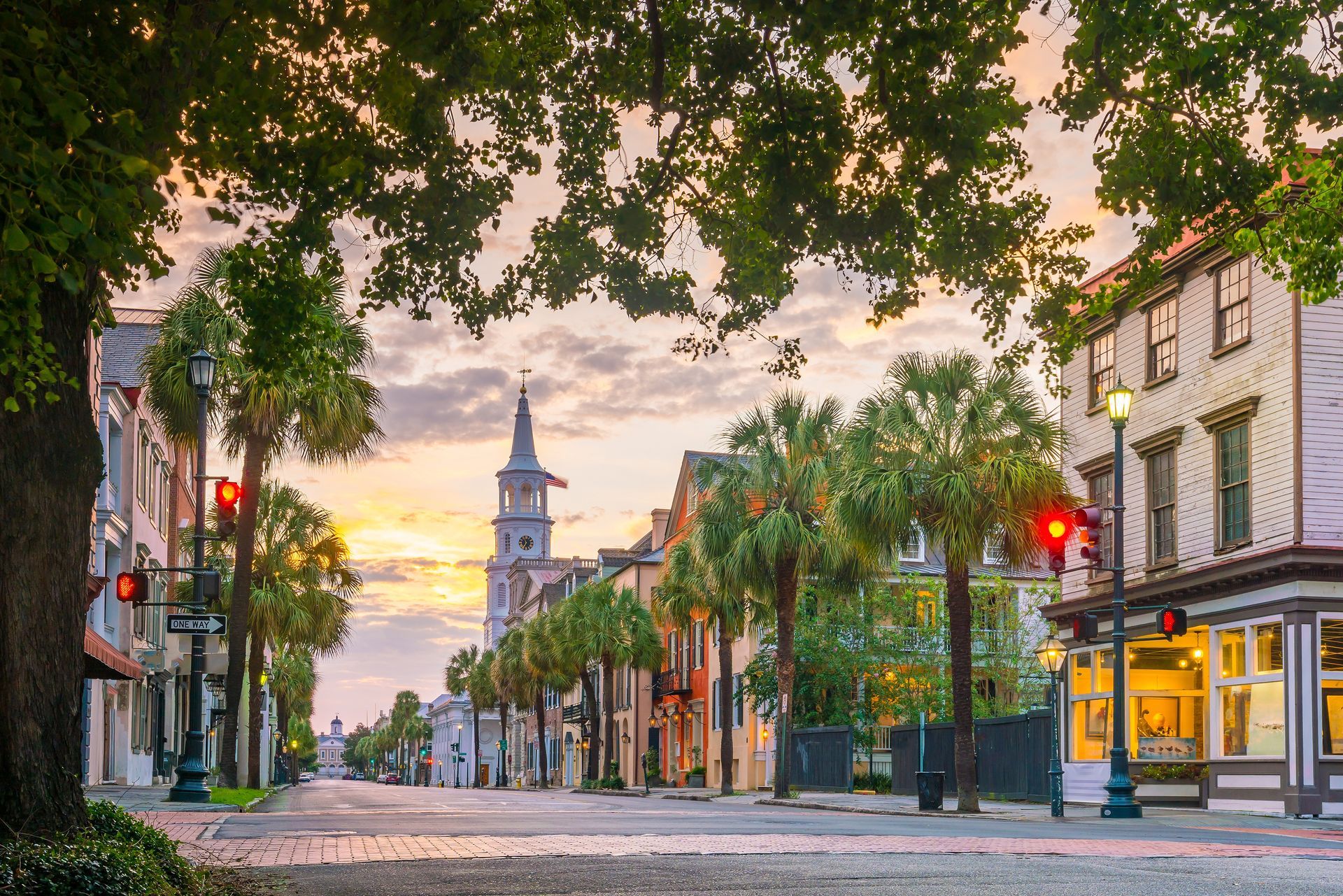 Street scene in Charleston, South Carolina, lined with buildings, palm trees, and a church steeple at dusk.