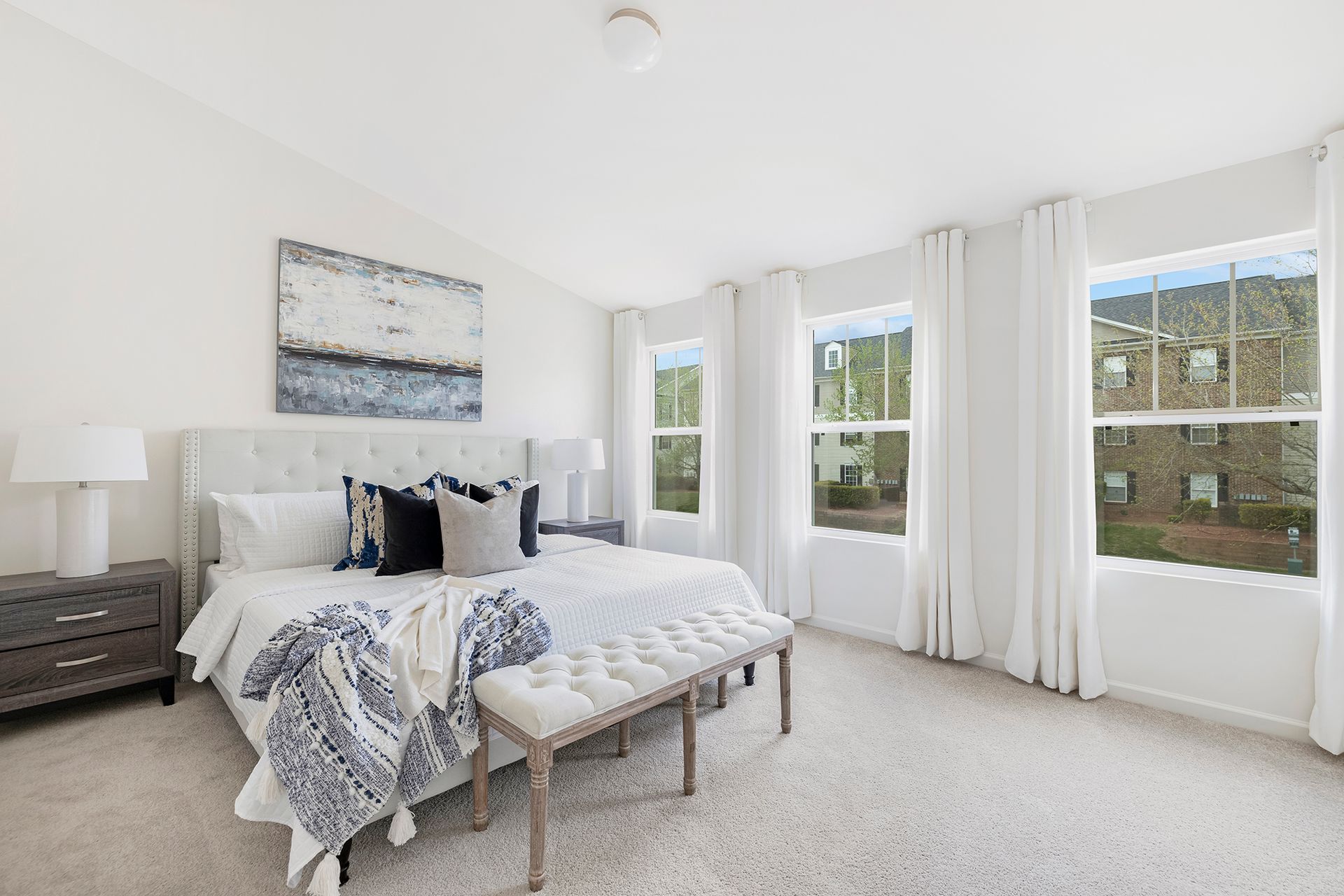 A bedroom with a king size bed , a bench , two windows and a painting on the wall.