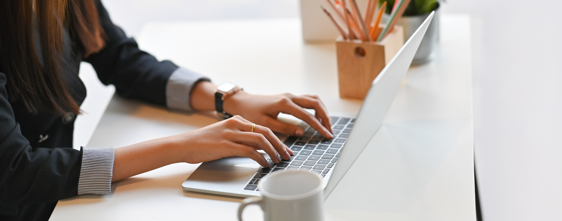 Person typing on a laptop at a white desk; mug and pencils present.