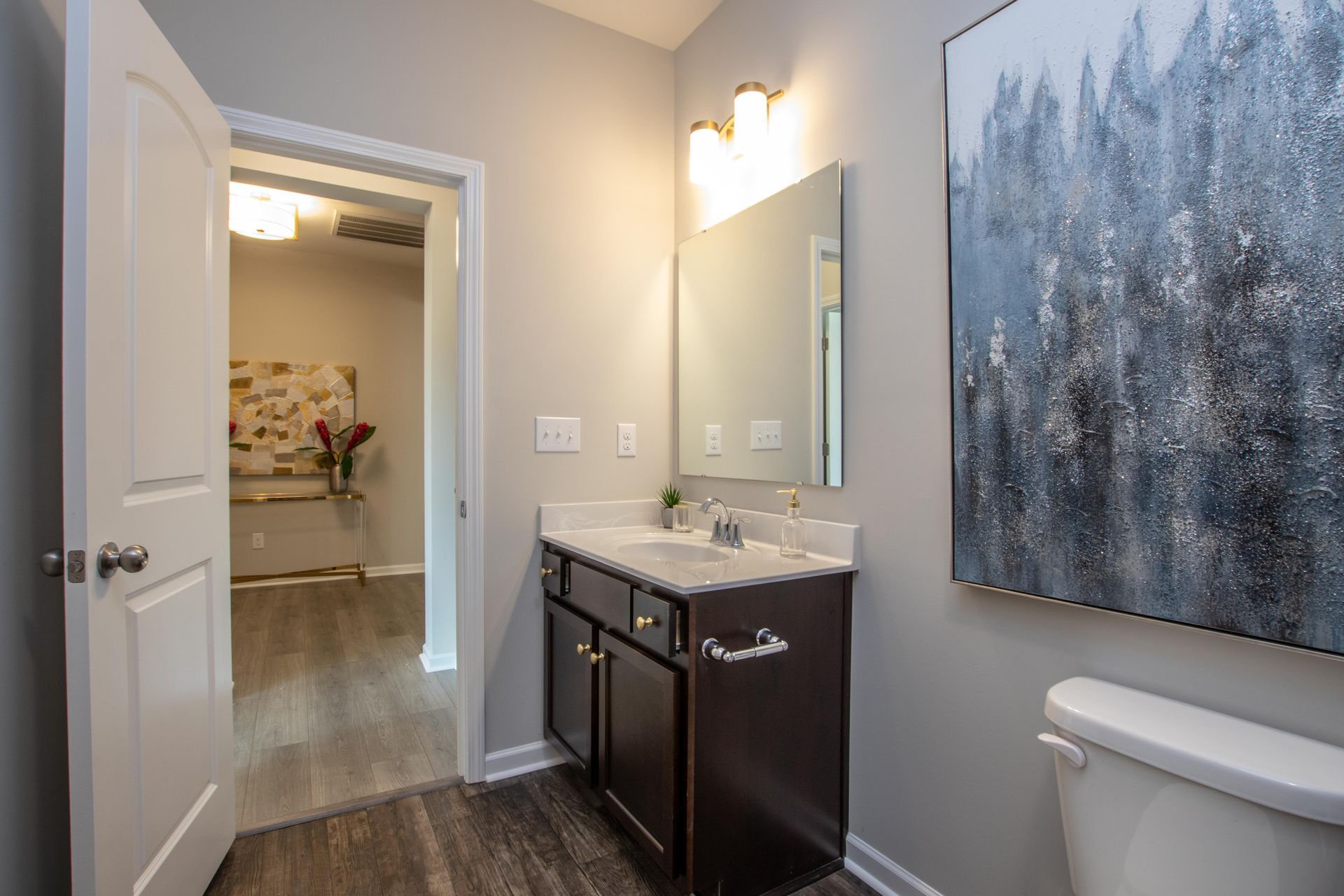 A bathroom with a sink , toilet , mirror and painting on the wall.