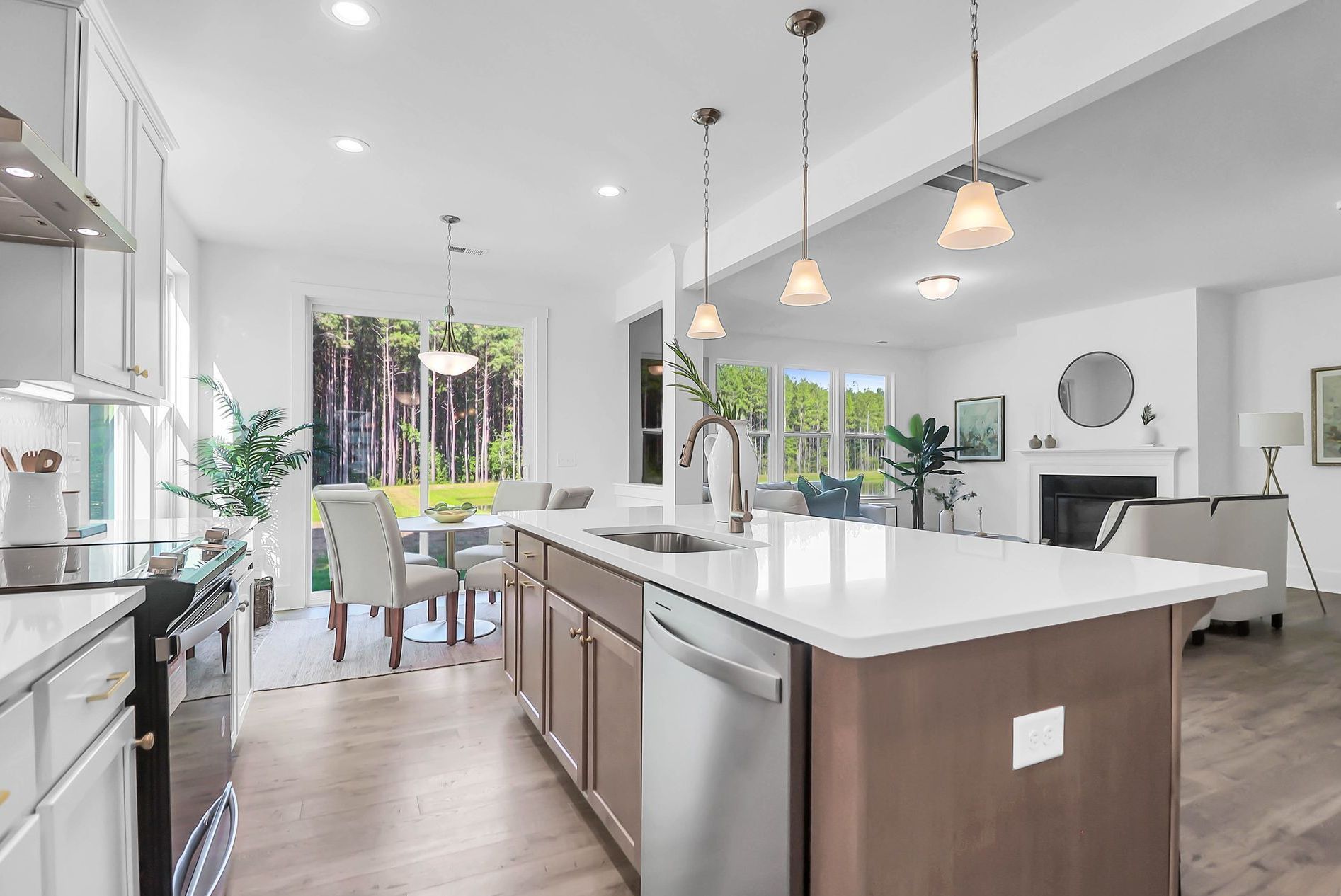 Open-concept kitchen with island, dining area, and living room, white and brown tones, overlooking a wooded view.