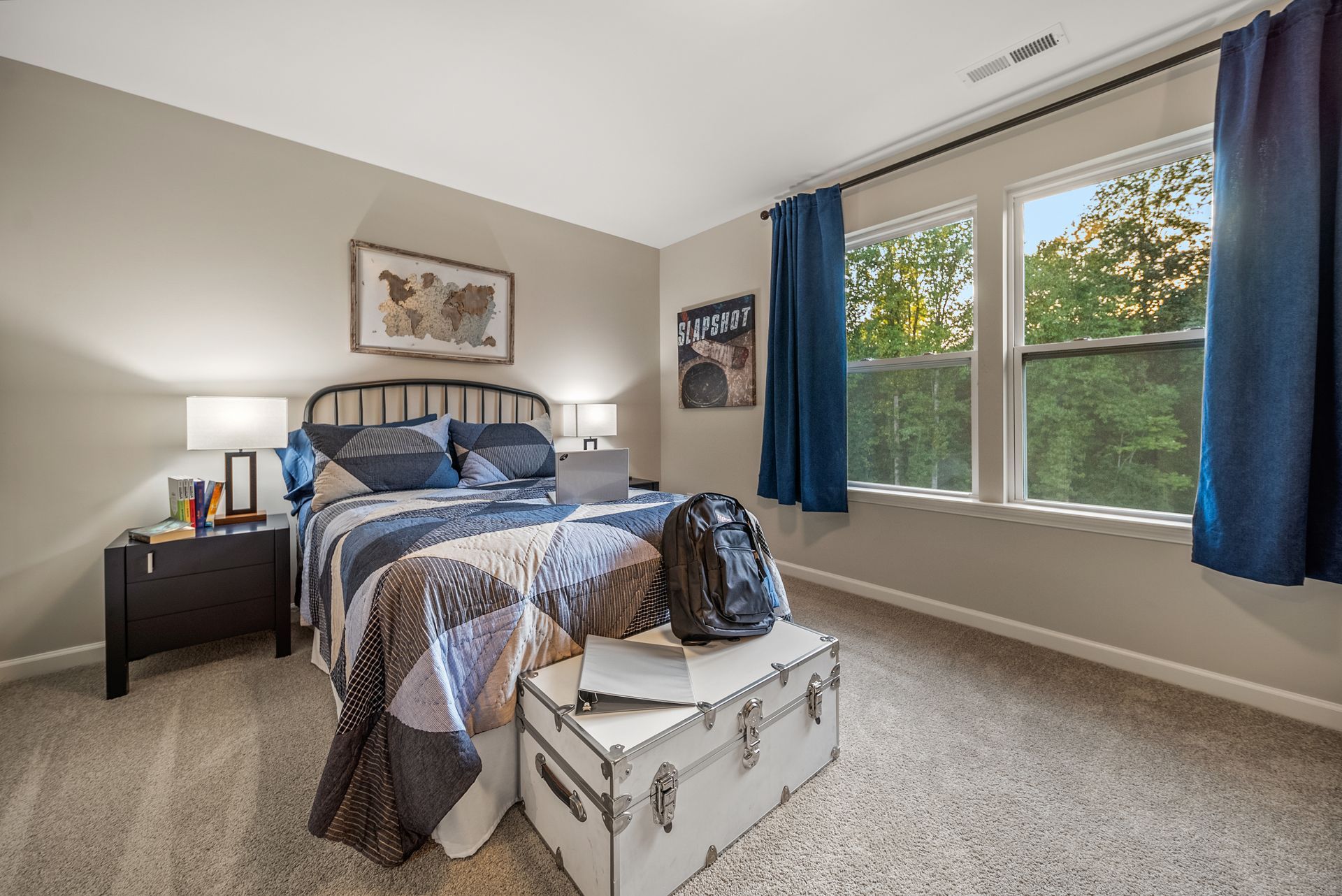 A bedroom with a bed , nightstand , trunk and two windows.