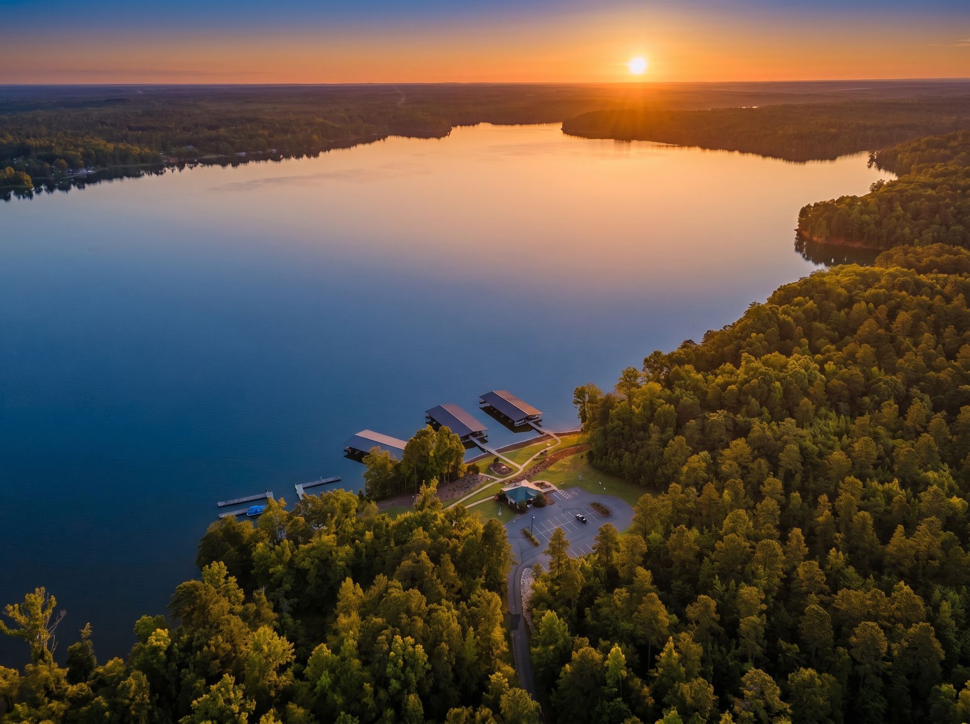 Sunset over a calm lake bordered by dense forest, with a small dock and buildings on the shore
