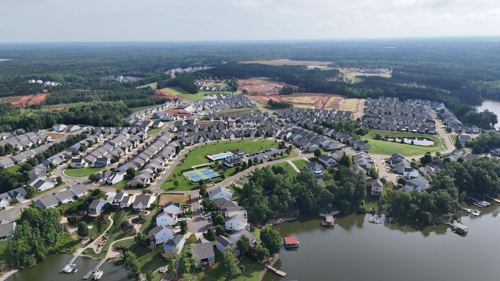 Aerial view of a lakeside residential neighborhood with houses, trees, and open space under a cloudy sky.