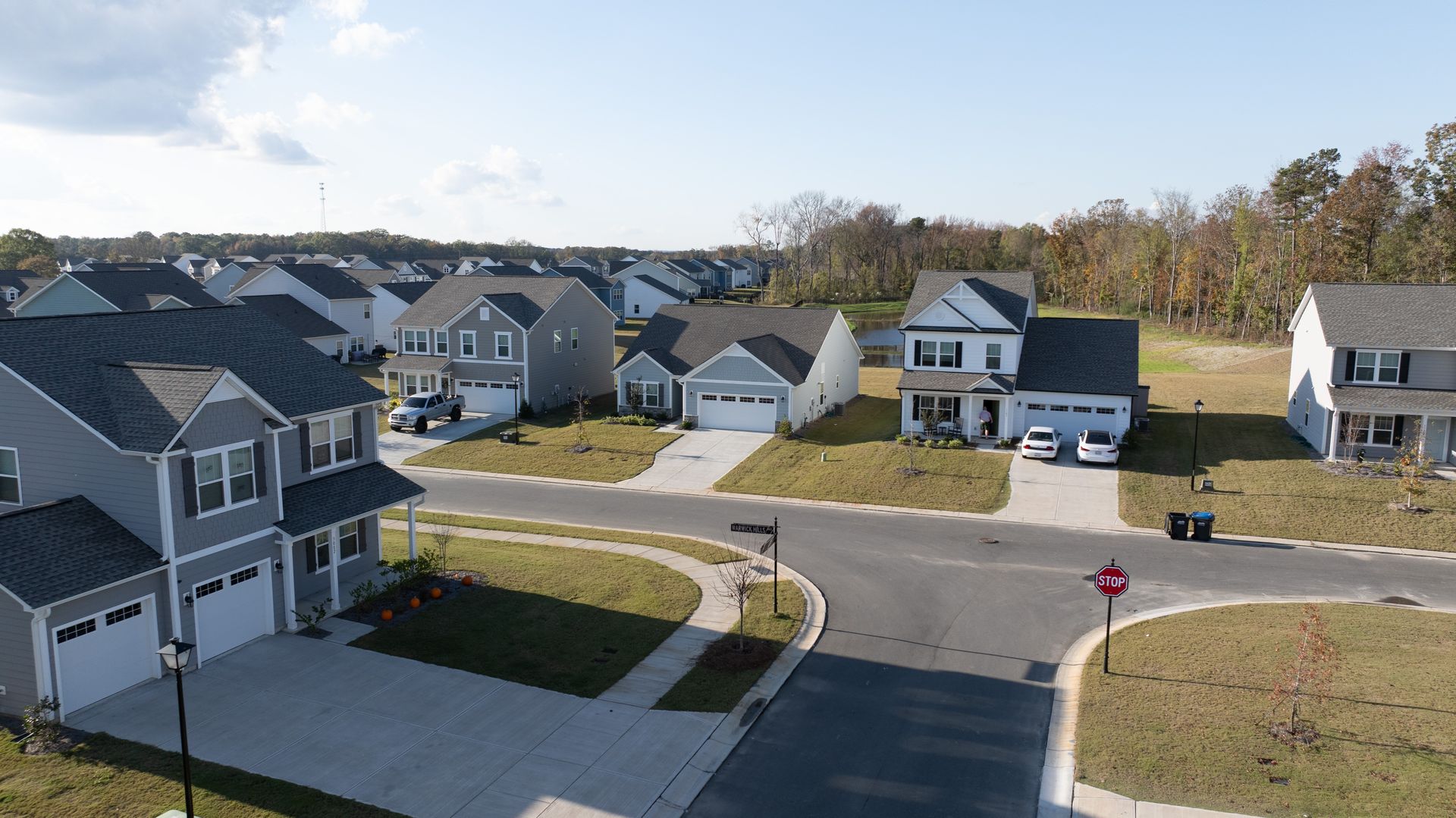 Suburban neighborhood with gray houses, driveways, trees, and a clear blue sky.