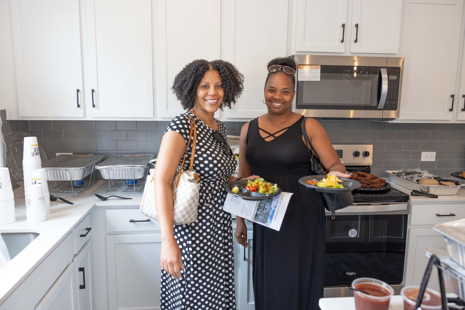 Two Black women smiling in a modern white kitchen; one holds food plates, the other a handbag.