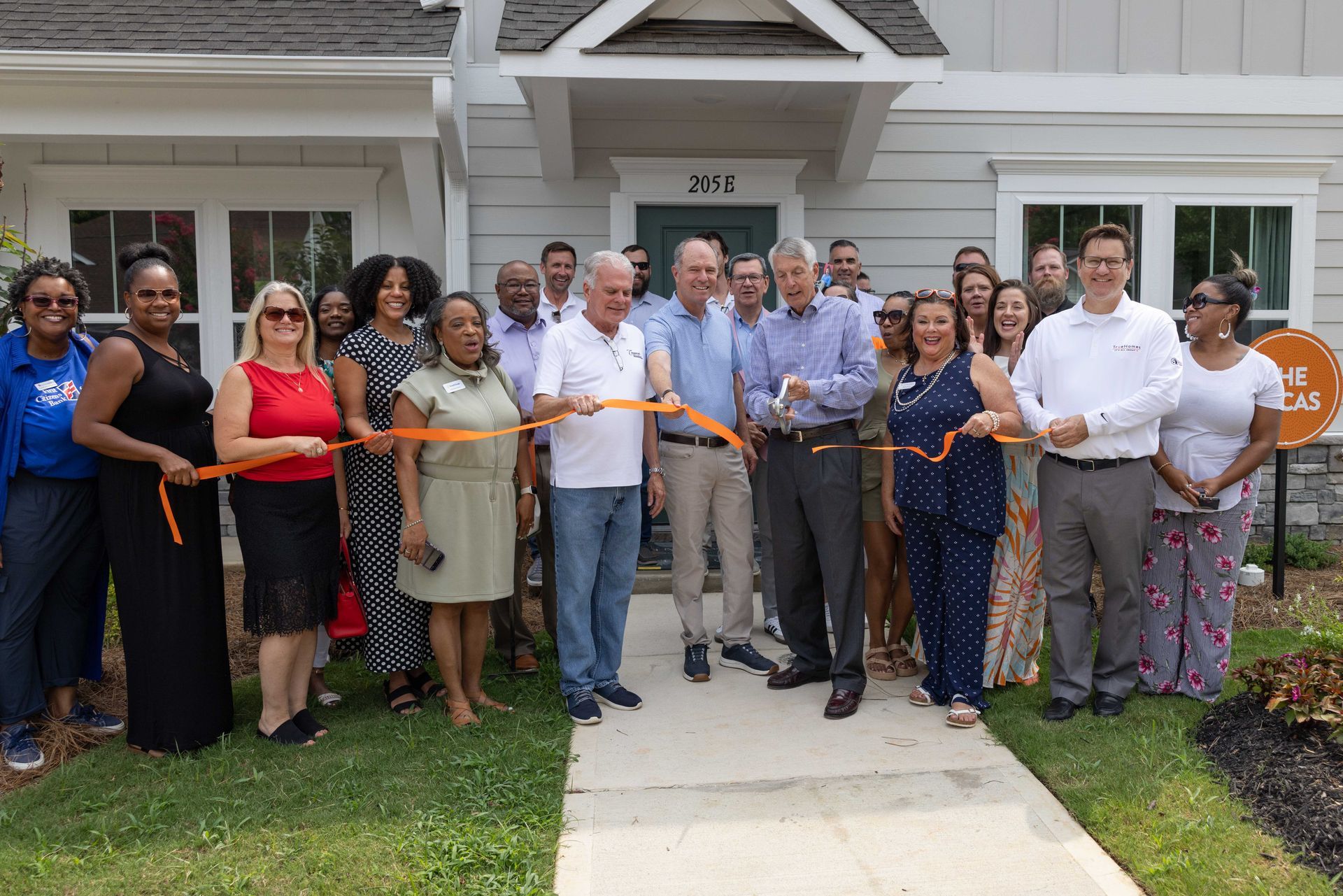 Group of people cutting an orange ribbon in front of a house, celebrating an opening.