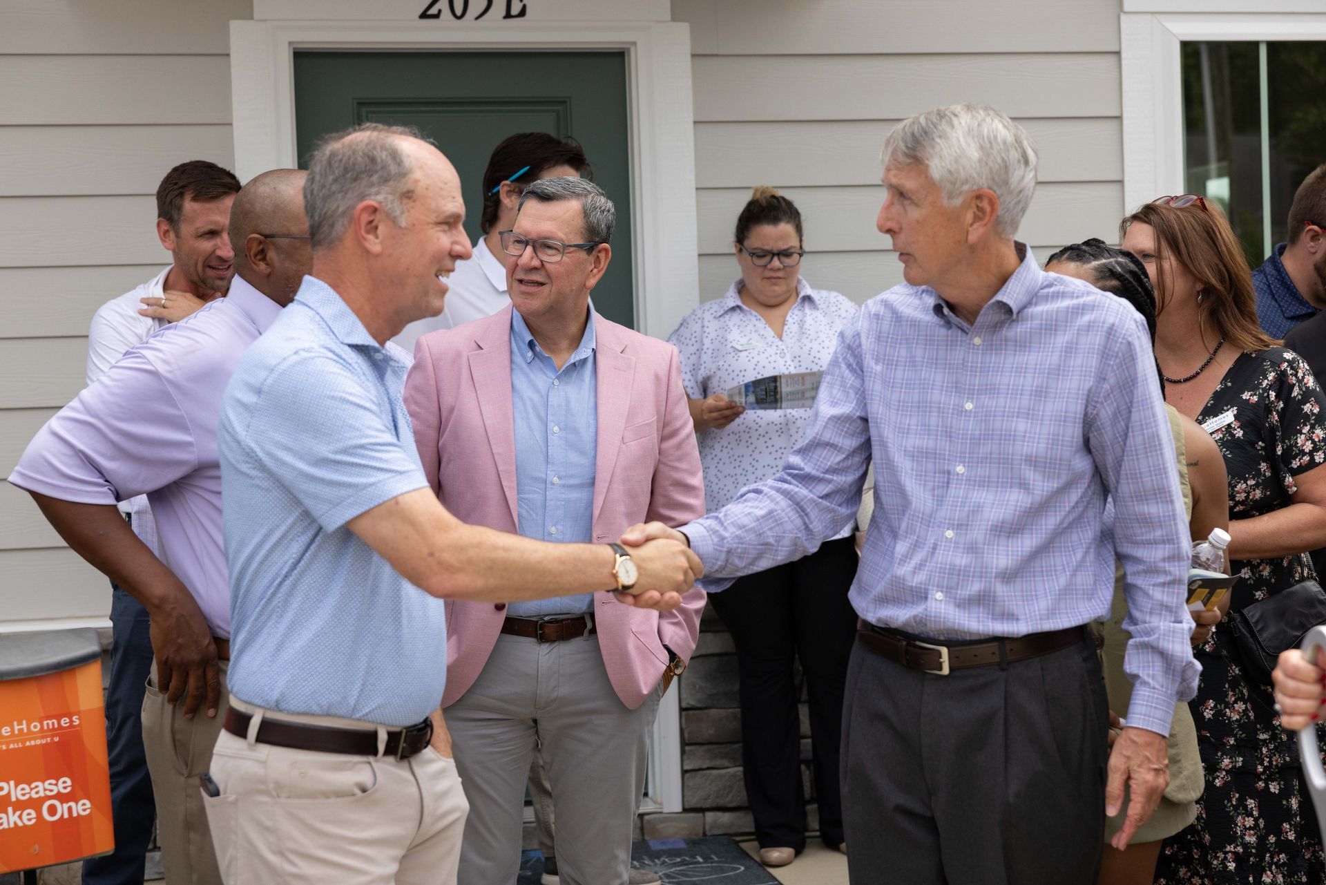 Two men shaking hands in front of a light-colored building; others watch.