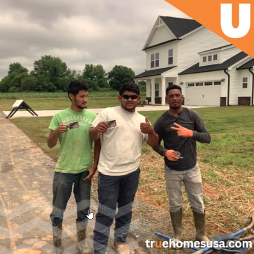 Three construction workers posing in front of a new white house. They are holding cards and giving thumbs-up.