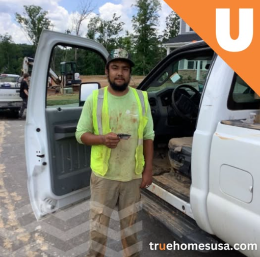 Construction worker stands by white truck; wears safety vest, cap, and holds a tool.