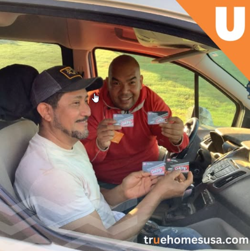 Two men in a car holding up gift cards, smiling. One wears a cap, the other a red shirt.