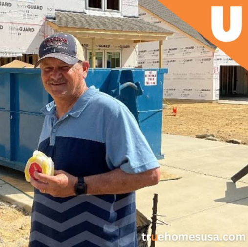 Man at construction site, eating. Wearing blue polo, baseball cap. Blue dumpster, unfinished house in background.