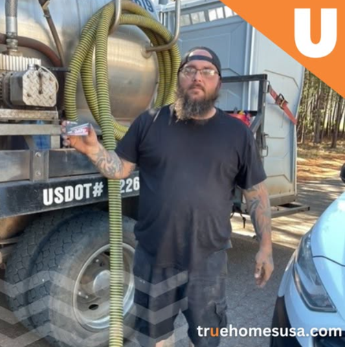 Man in front of a septic tank truck holding something, with tattoos, wearing black, outdoors.