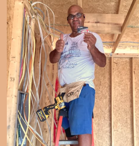 Electrician on ladder with tool, thumbs up, wiring in unfinished structure.