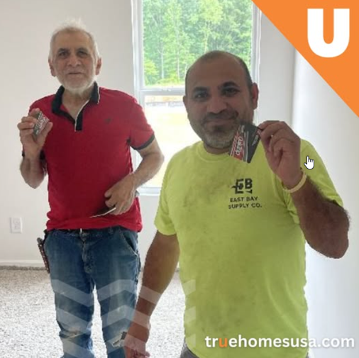 Two men holding cards, smiling, inside a bright room; one in red, the other yellow.