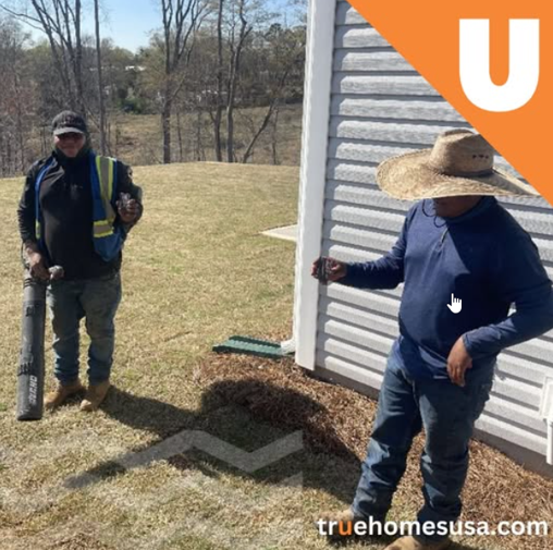 Two workers, one with leaf blower, near a house with blue siding on a sunny day.