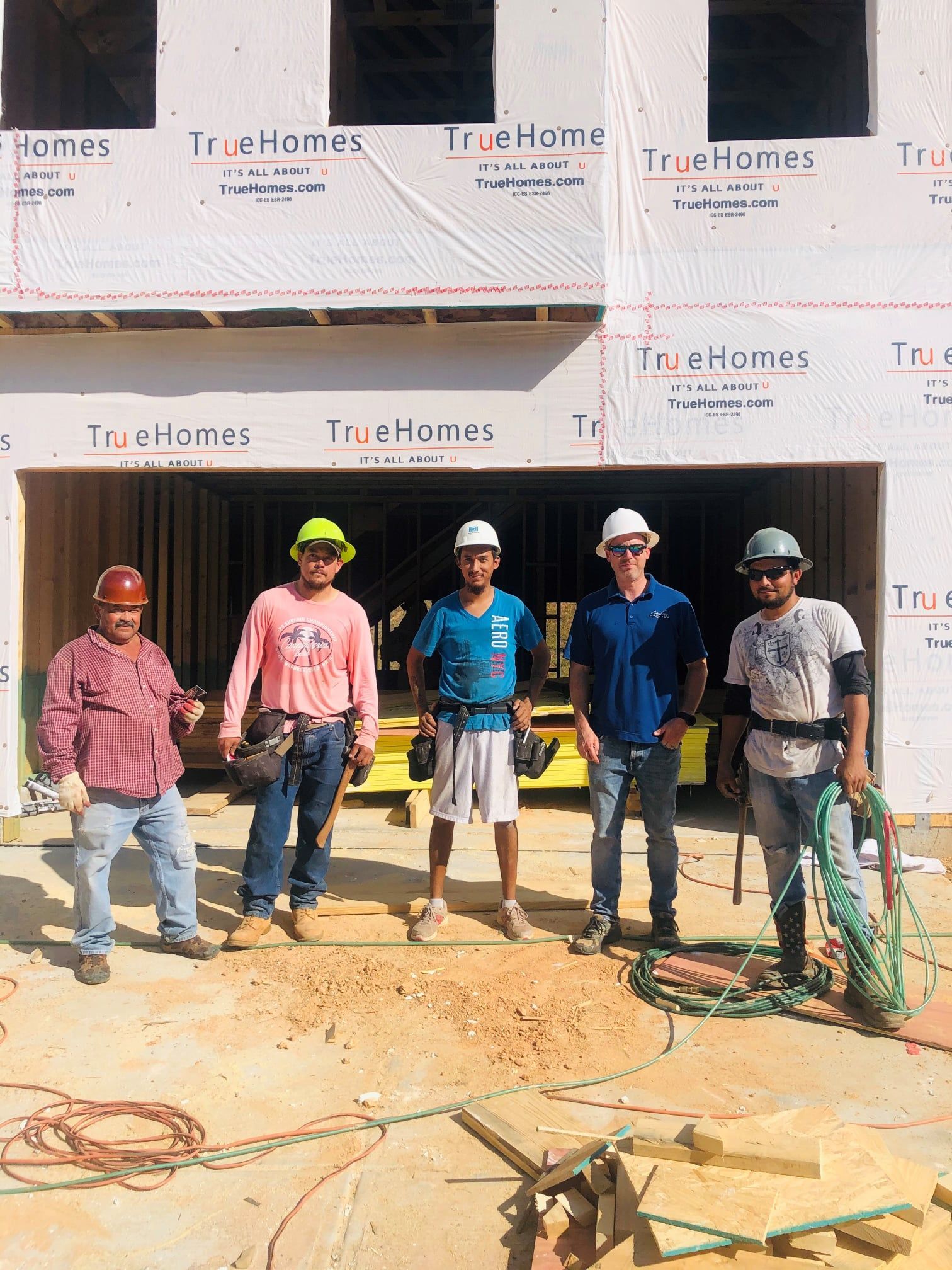 Five construction workers standing in front of a new house, wearing hard hats, tools visible, sunny day.
