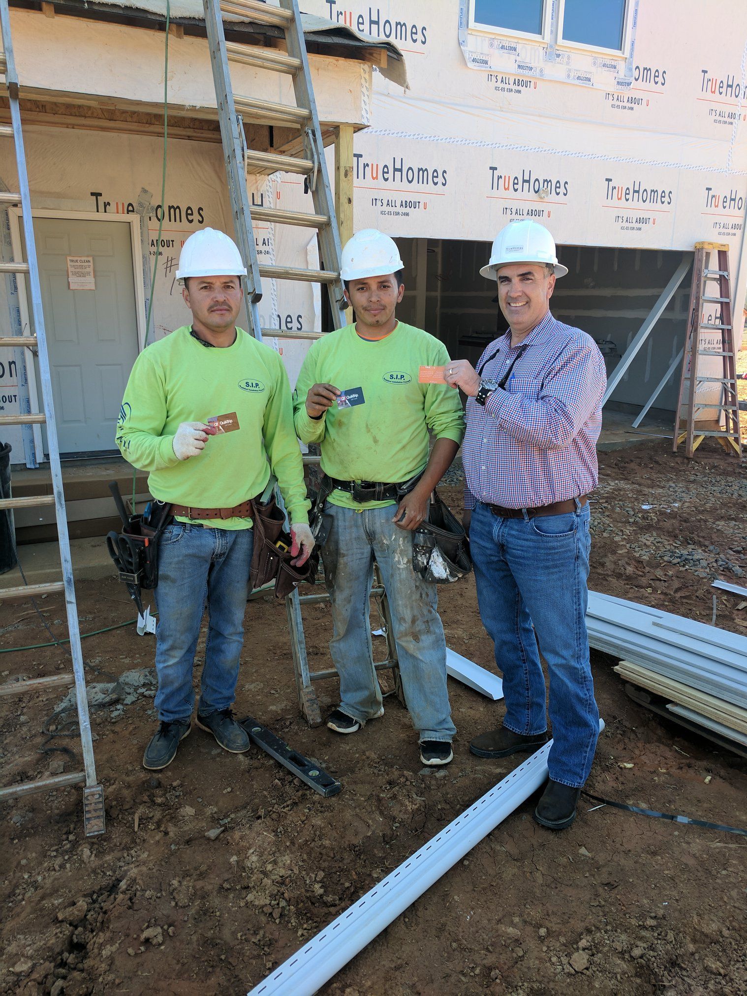 Three construction workers, one giving a thumbs up, pose in front of a house under construction.