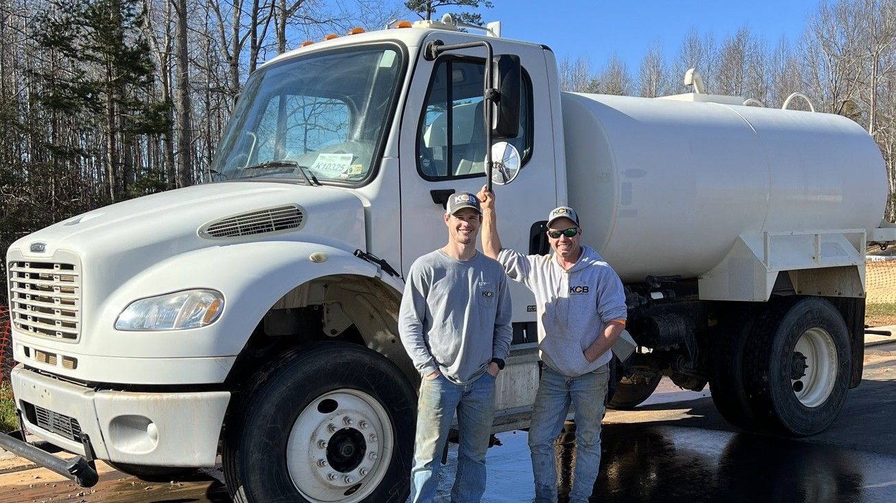 Two men stand beside a white water tanker truck in front of trees.