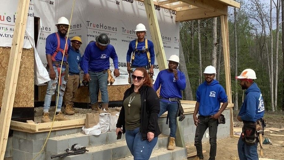 Construction workers in blue and a woman in black on a building site, smiling and posing.