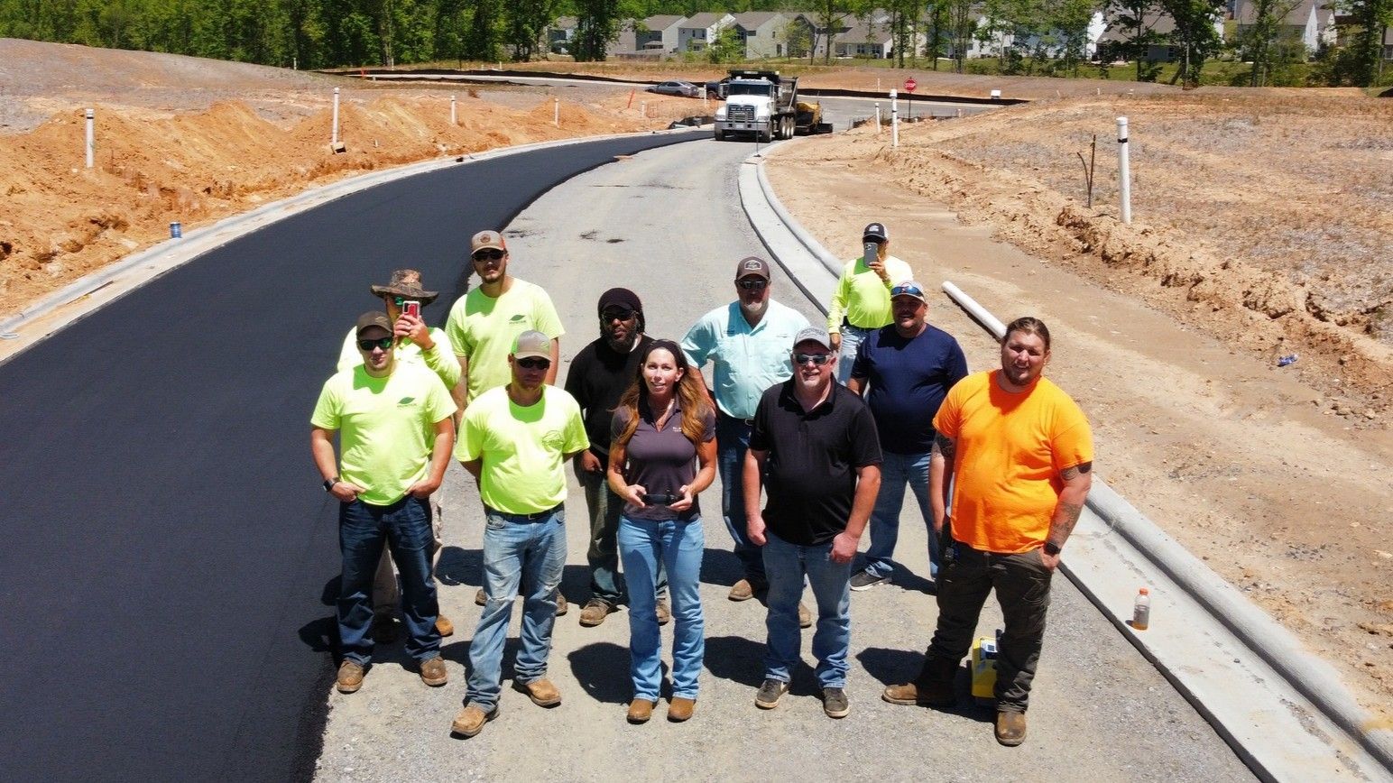 Construction crew posing on a newly paved road; sunny day, dirt and trees in background.