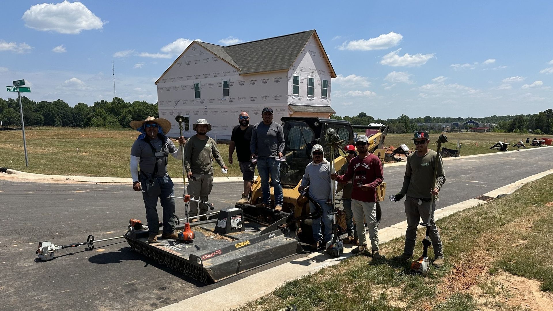 Construction crew with equipment on a paved road in front of a new house, sunny day.
