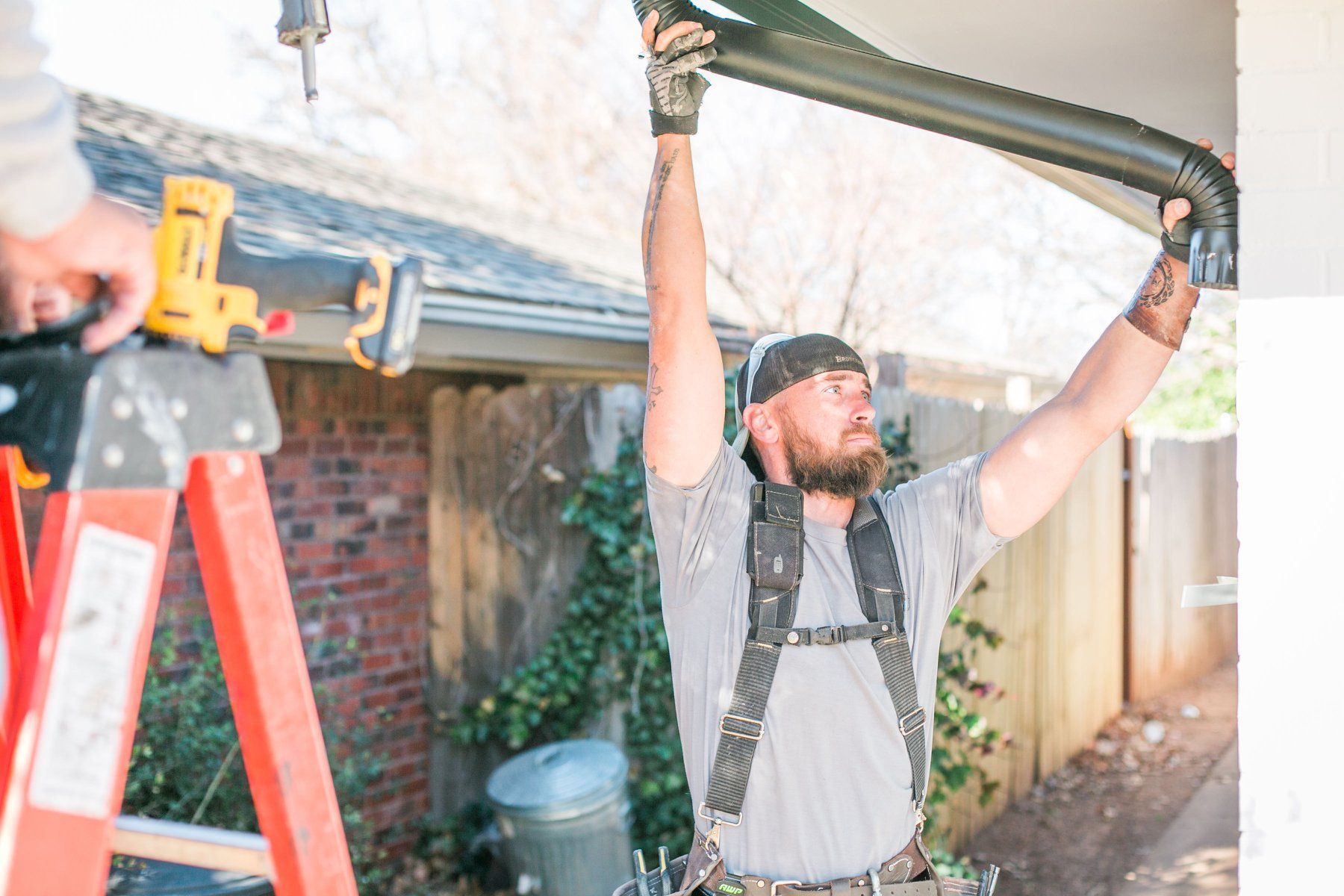 A man is hanging from a ladder while installing a gutter on a roof.