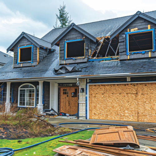 Damaged house with boarded-up windows and missing roof sections, debris on the lawn; grey sky.