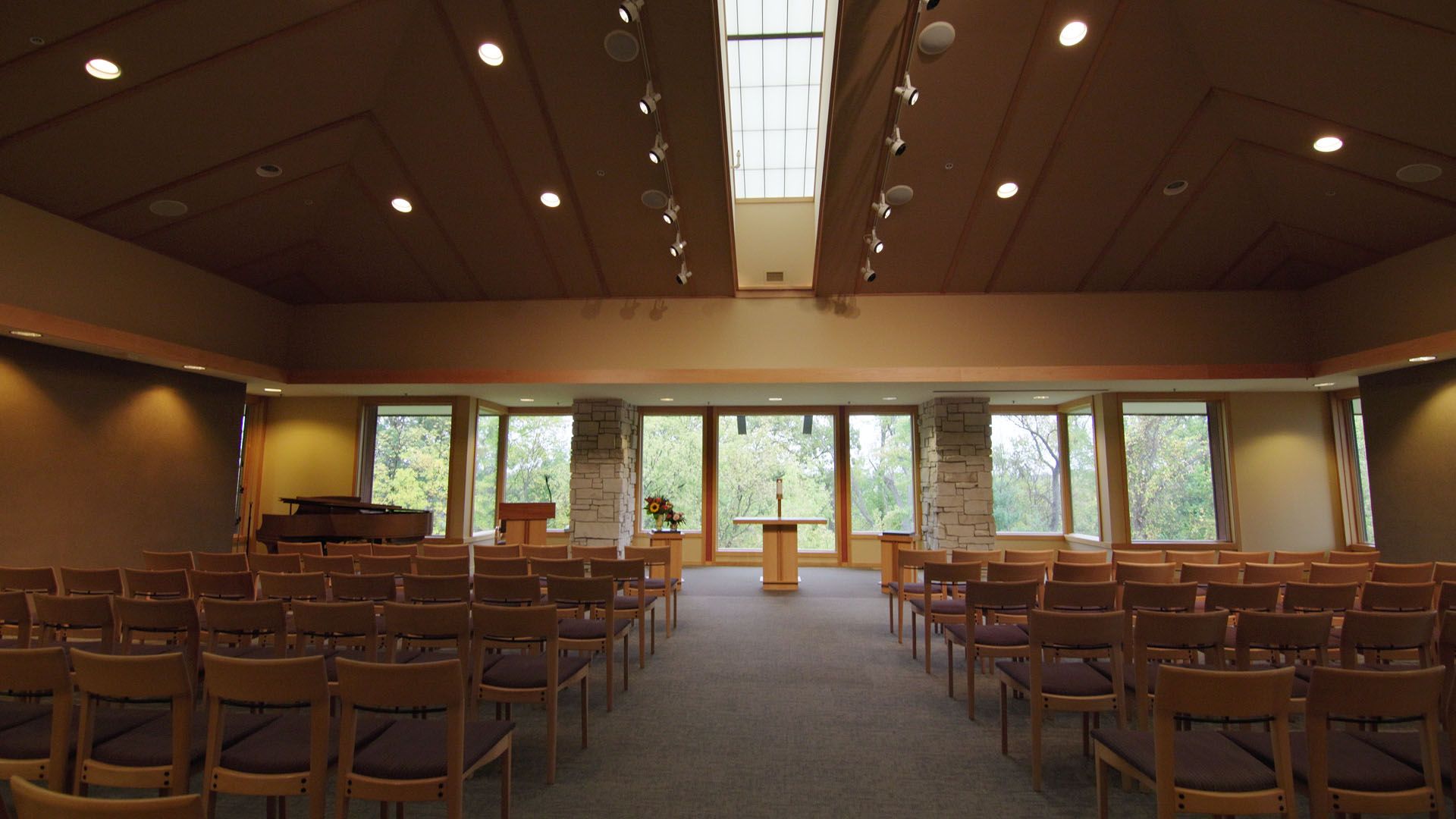 Interior of a chapel with rows of chairs, a pulpit, and windows overlooking greenery.