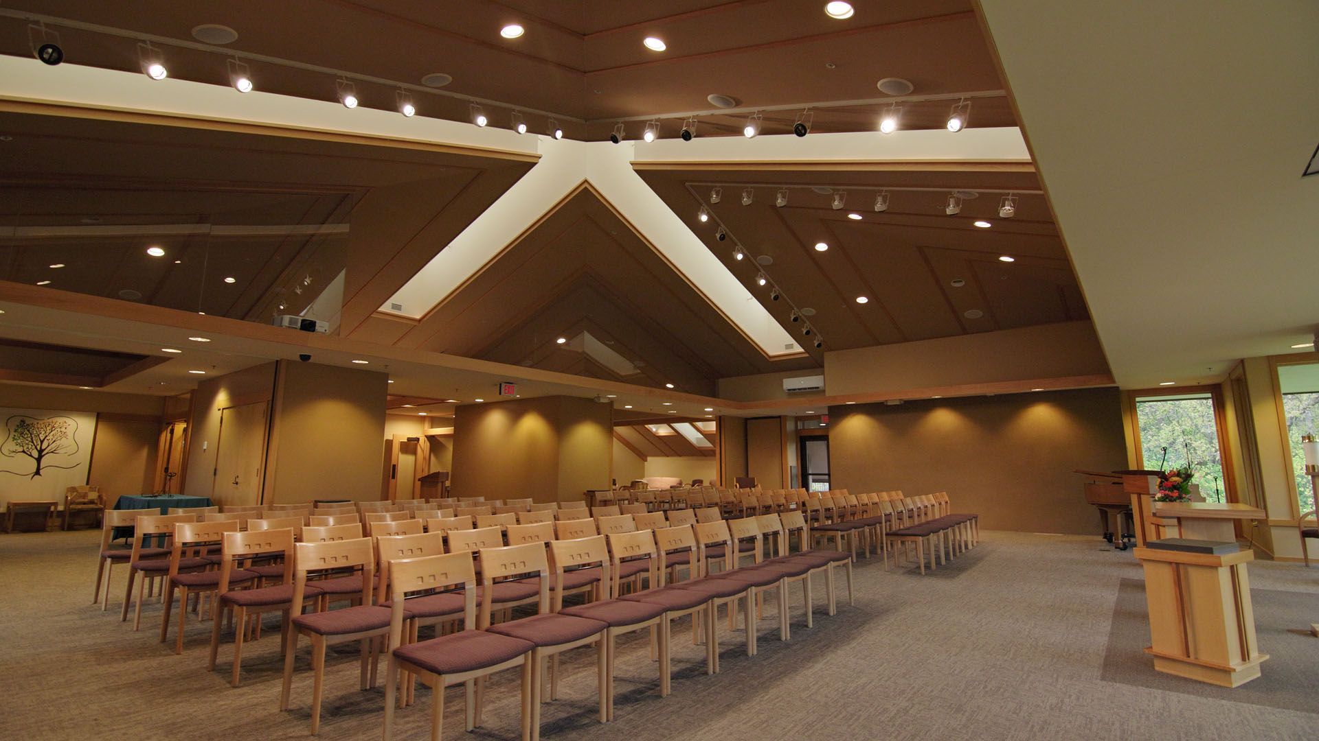 Rows of wooden chairs face a podium in a light-filled interior with a sloped ceiling and recessed lighting.