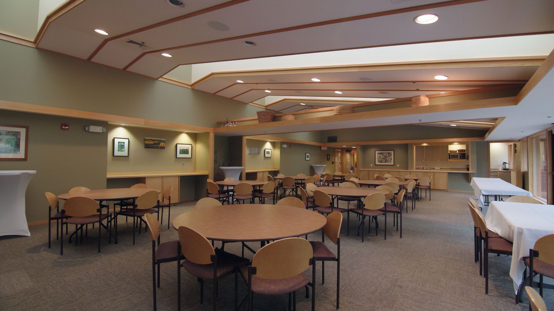 A dining room with round tables and chairs. Beige walls, wood beams, and neutral carpet.