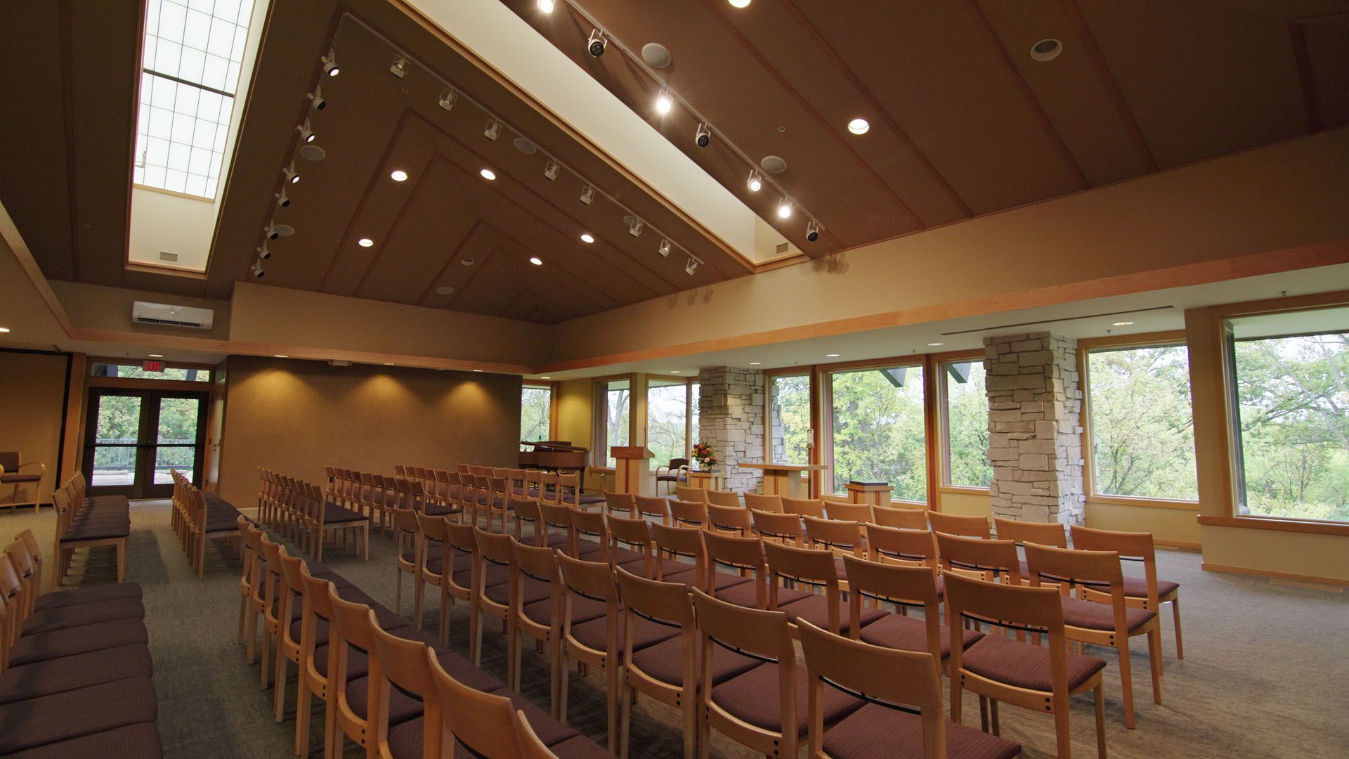 Rows of chairs face a podium in a chapel-like room with large windows, skylights, and warm wood tones.