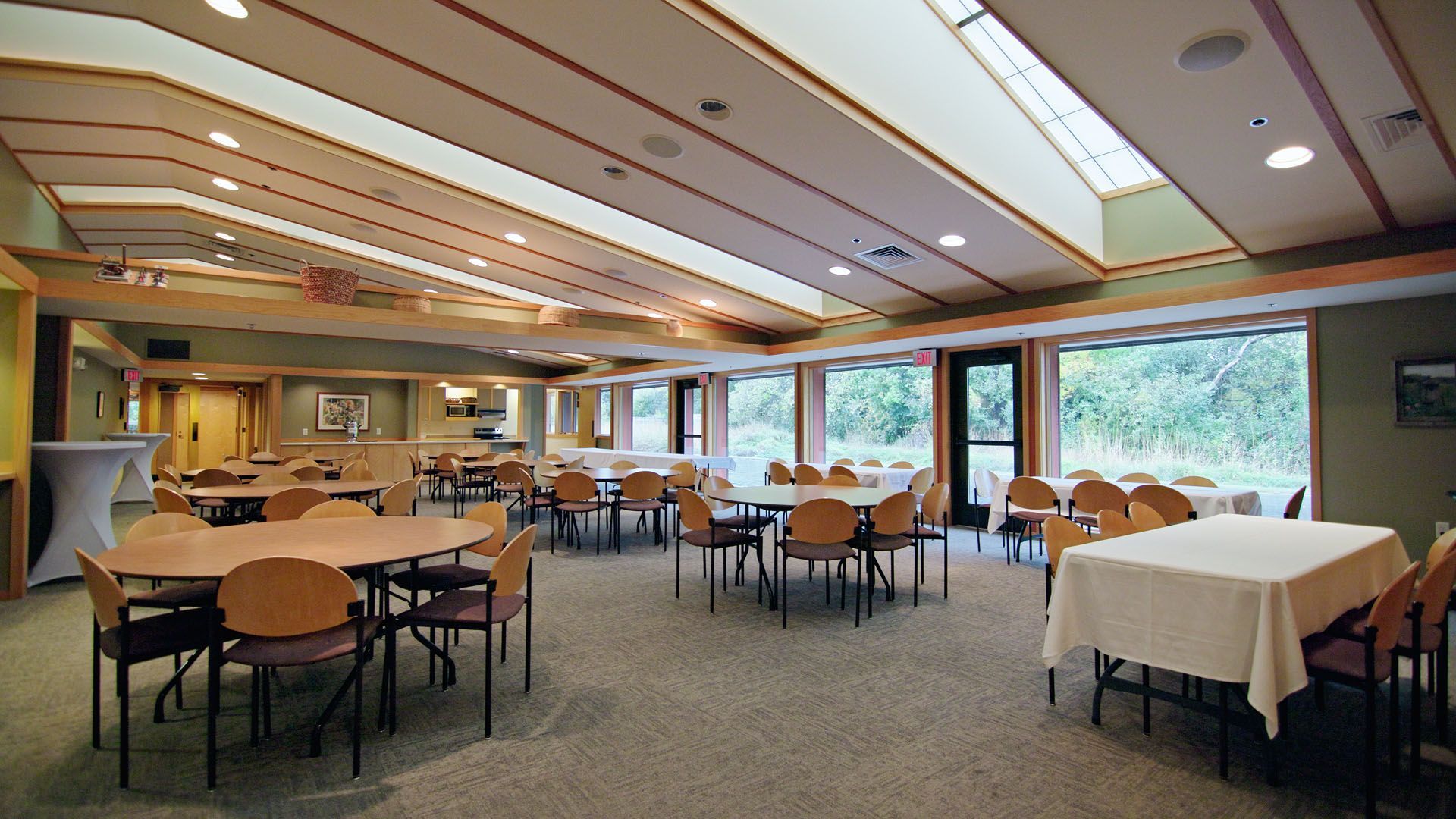 A bright, open room with round and rectangular tables, chairs, and large windows overlooking trees.