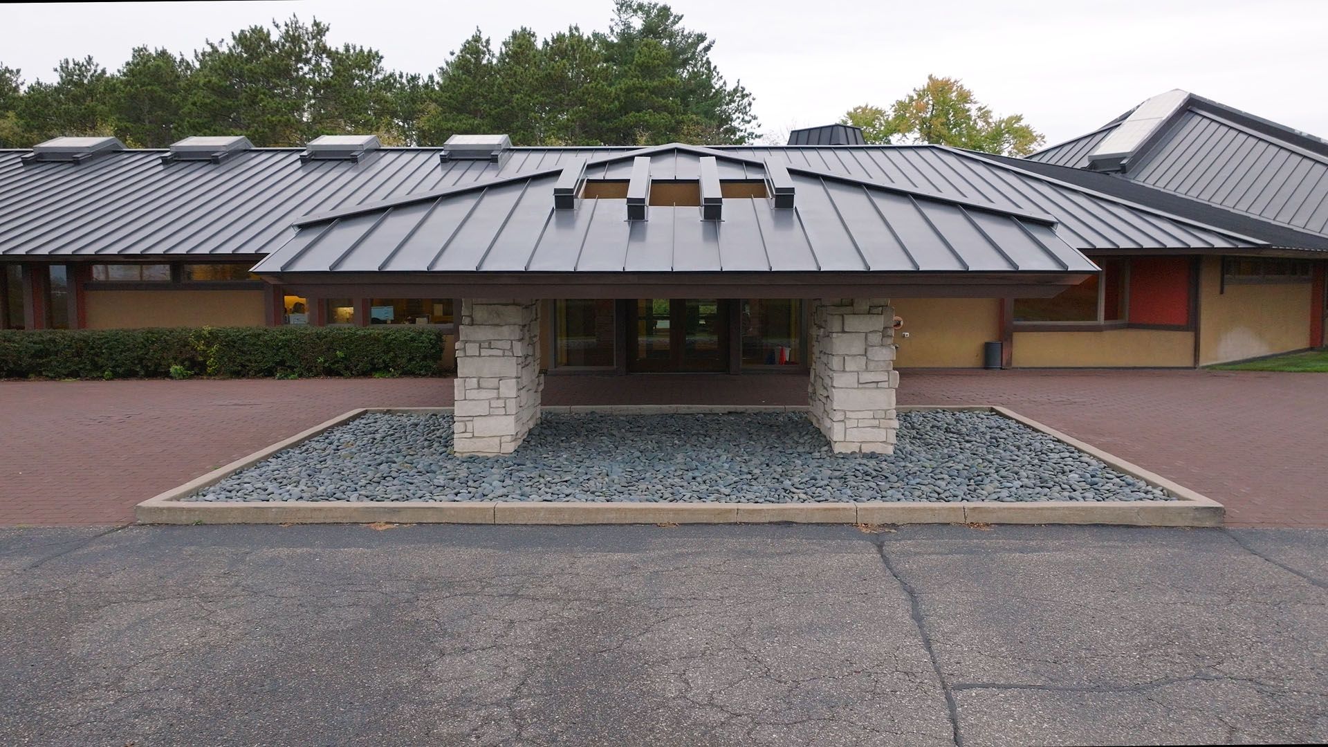 Building entrance with dark metal roof supported by stone pillars; gravel bed.
