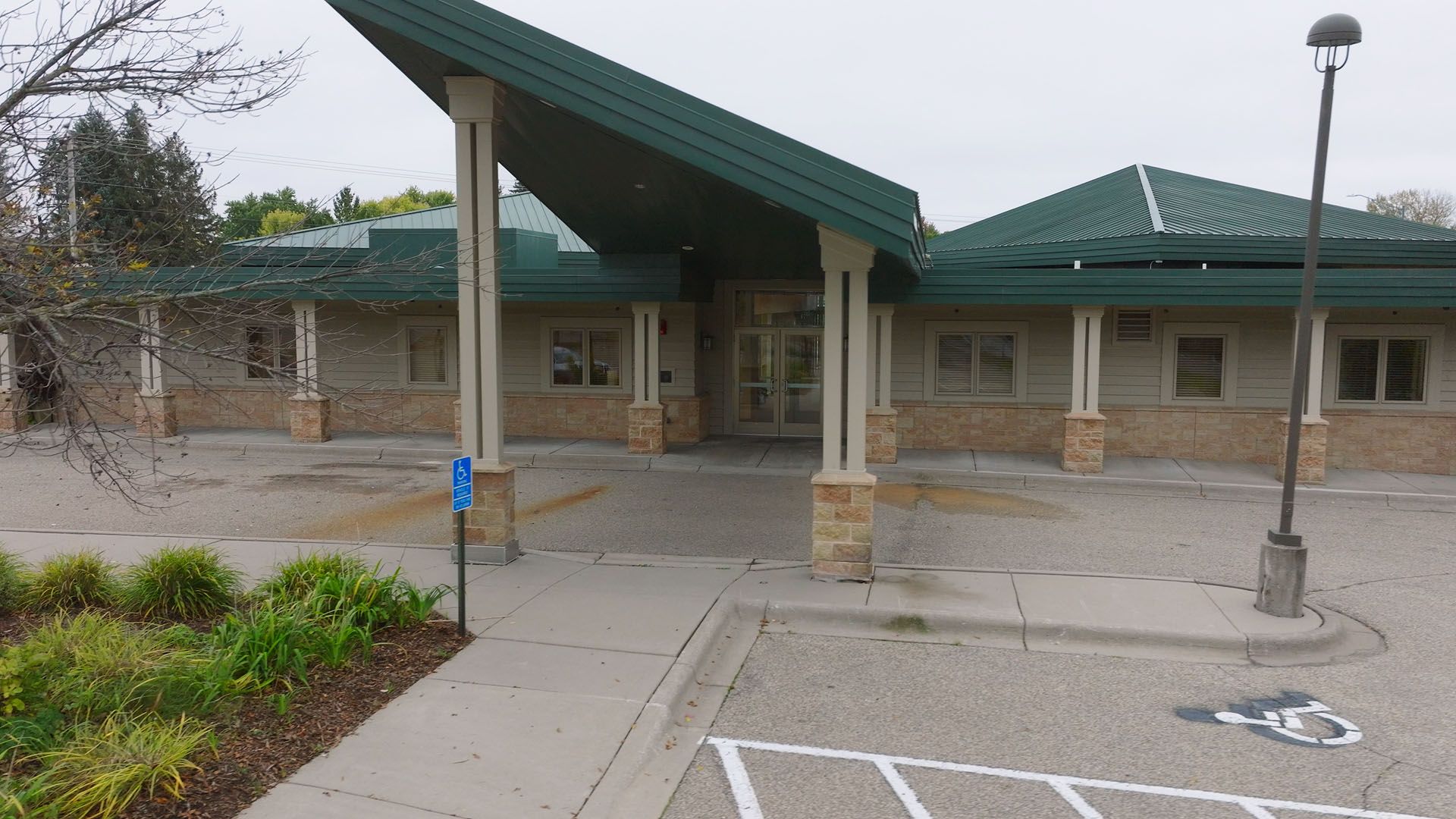 Building with green roof, light tan walls, and a covered entrance. Paved parking area with accessible parking sign.
