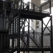 Escalera de metal negro con rellanos y barandillas de malla en entorno industrial.