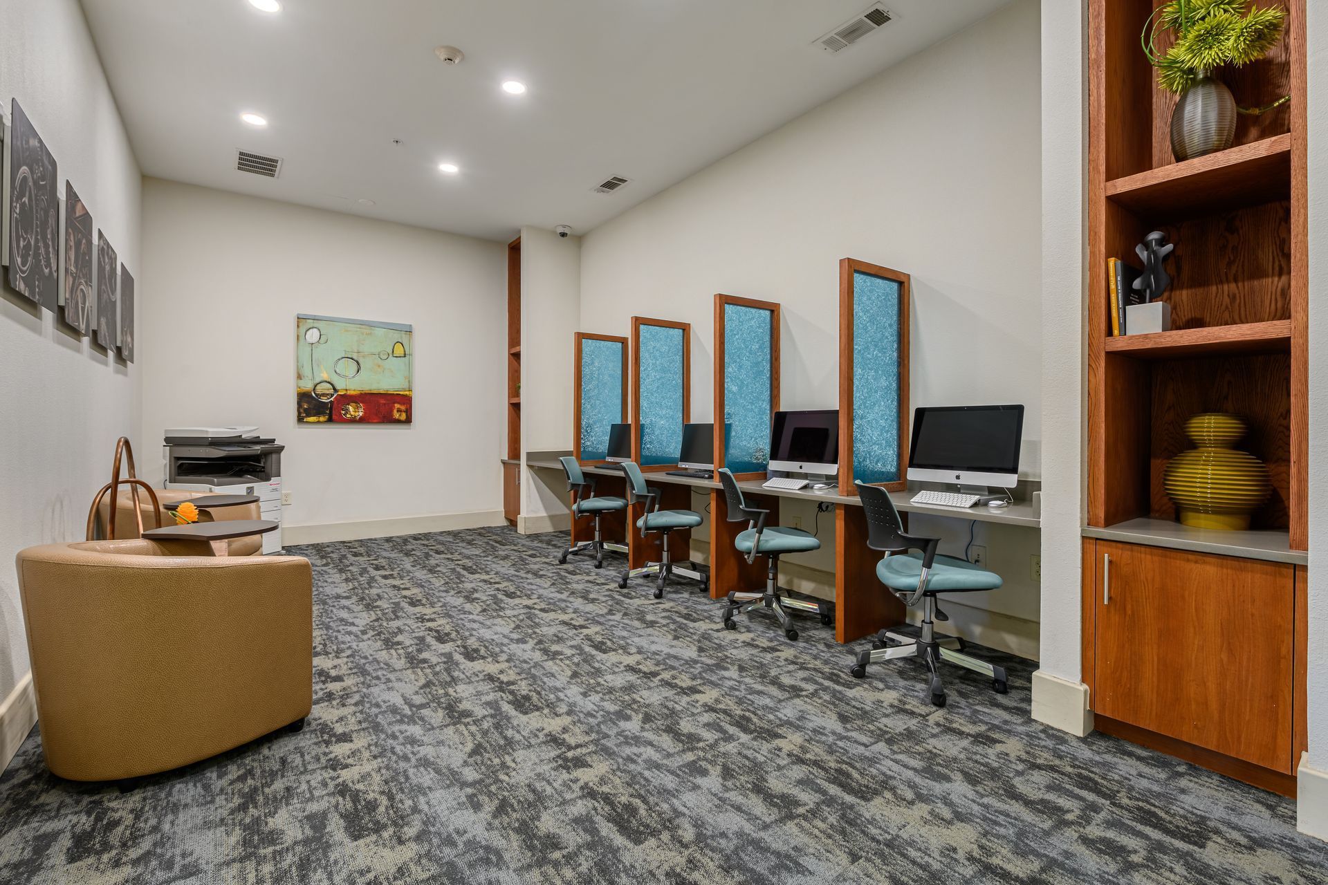 A business center with four computer workstations, a printer, a lounge chair, and a wooden shelf in a quiet room.