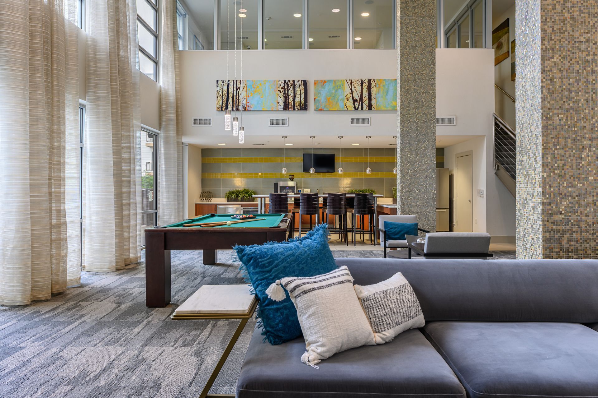 Modern apartment complex lobby with a pool table, bar seating, patterned carpet, and a large grey sofa with accent pillows.