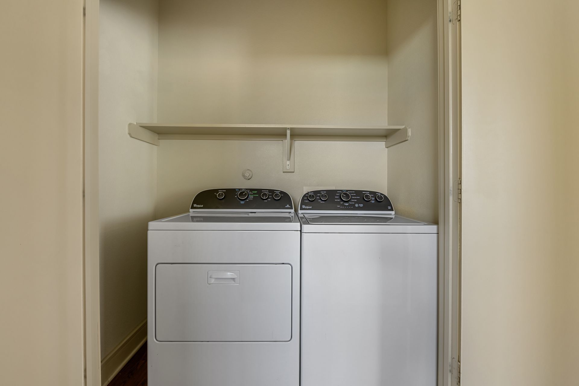 A white side-by-side washer and dryer set in a small, beige indoor laundry closet with a shelf above.