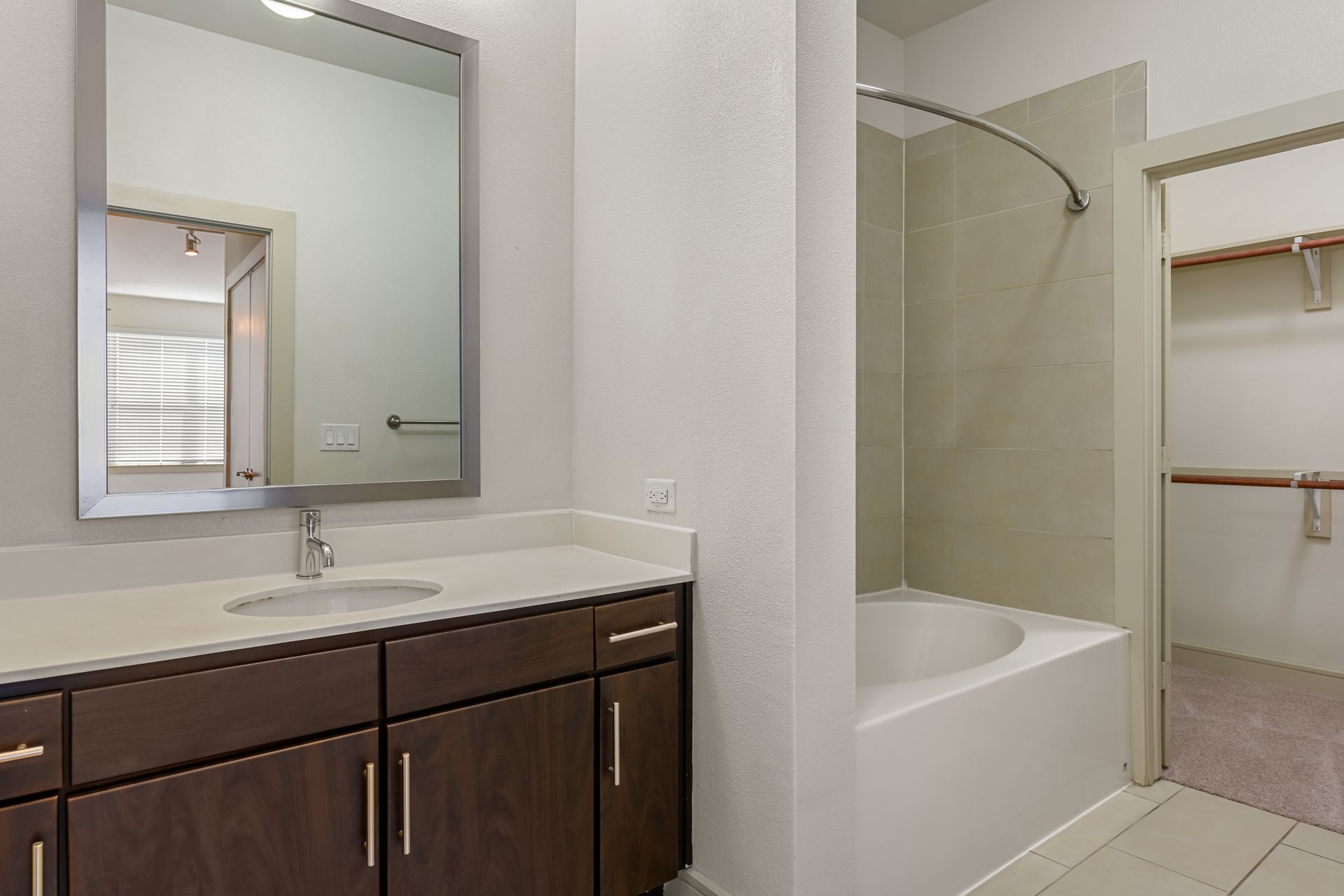 A bathroom vanity with dark wood cabinets, a mirror, and a tiled tub-shower combo next to a walk-in closet.