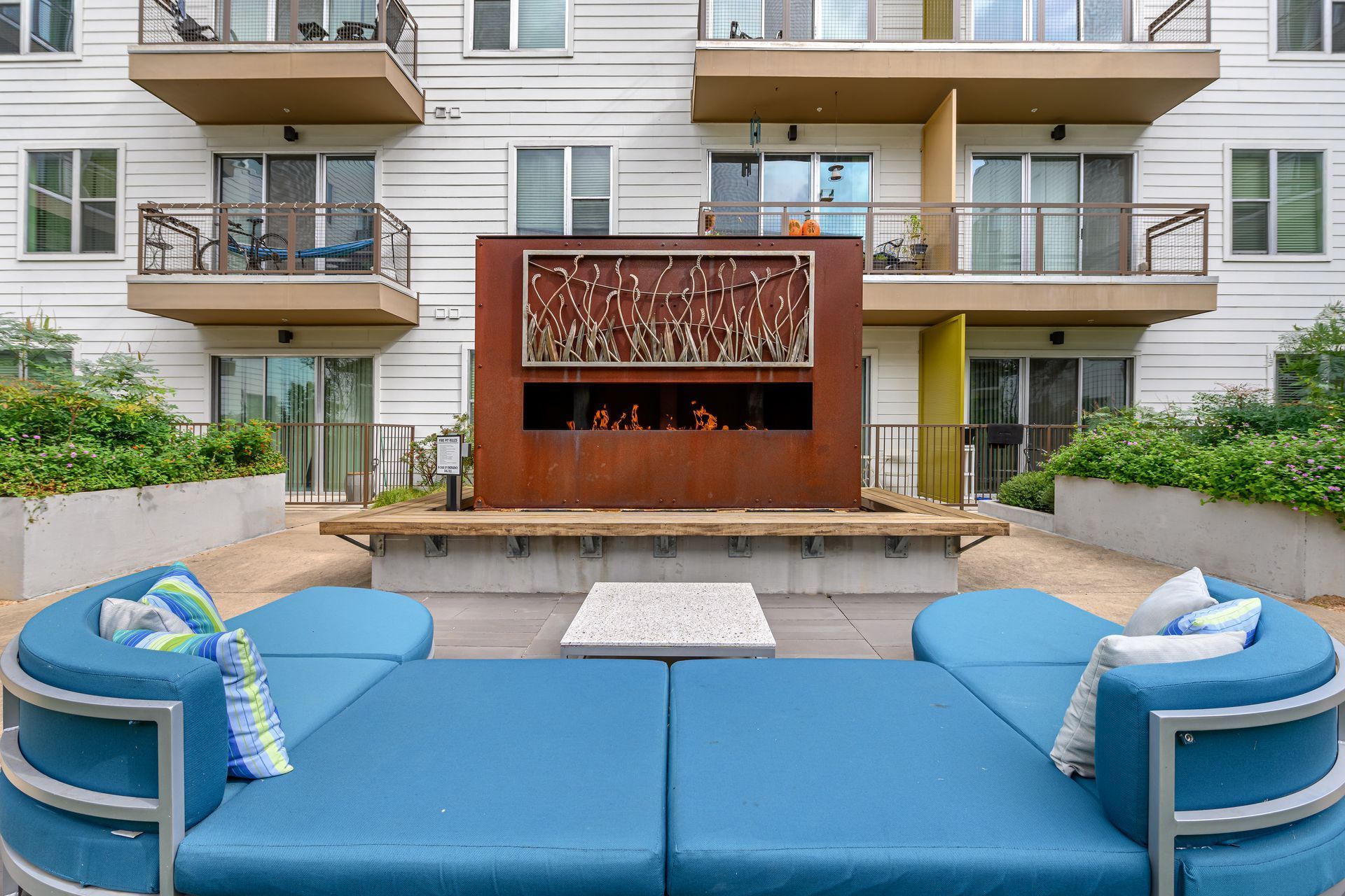 A large, curved blue sofa faces an outdoor fireplace on a patio in front of a multi-story apartment building.