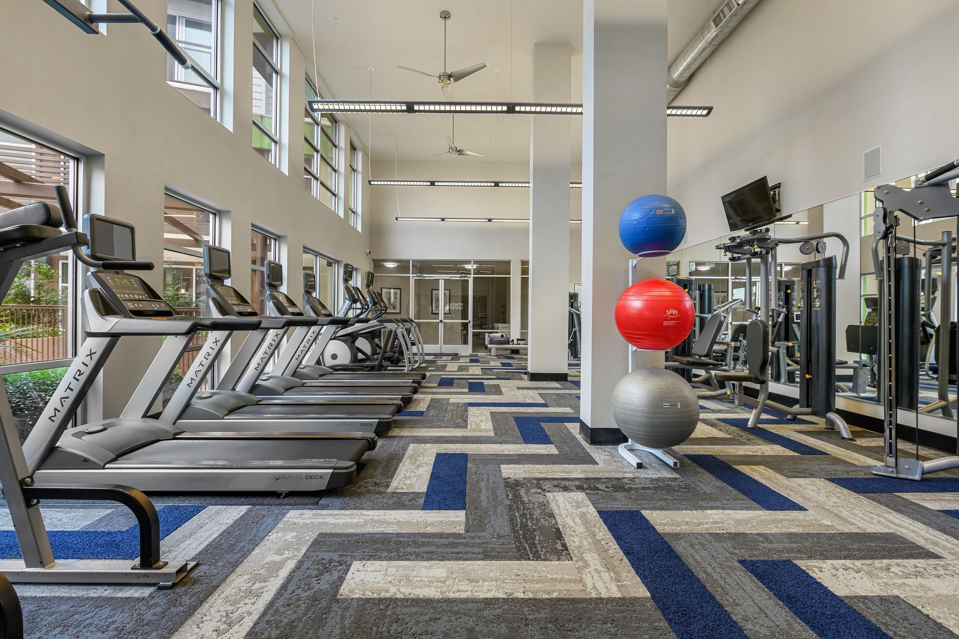 A bright fitness center featuring a row of treadmills, exercise balls on a rack, and patterned carpet flooring.