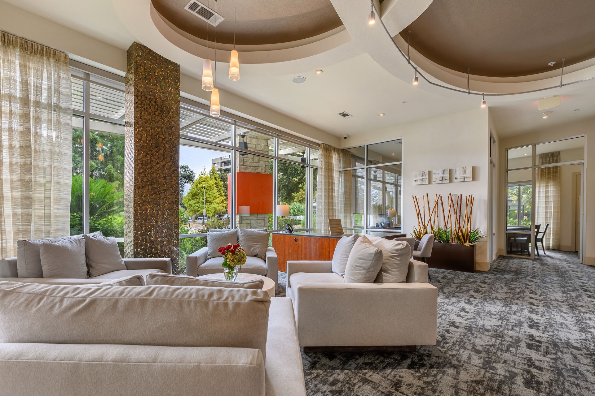 A modern lounge featuring light-colored sofas, patterned carpet, tall windows, and a circular recessed ceiling.