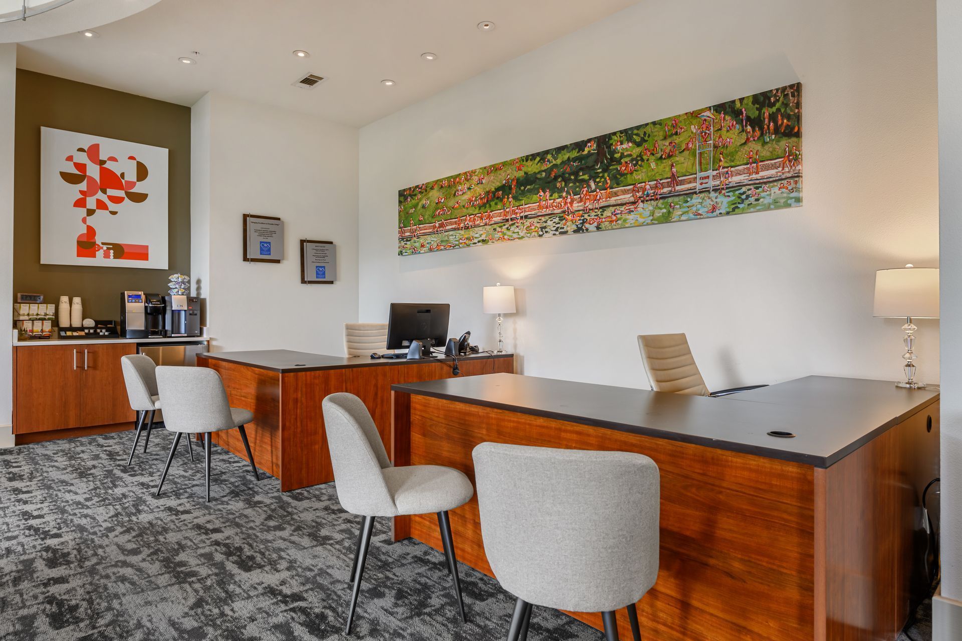 A modern office lobby with two wooden desks, gray chairs, a coffee station, and abstract wall art on a carpeted floor.