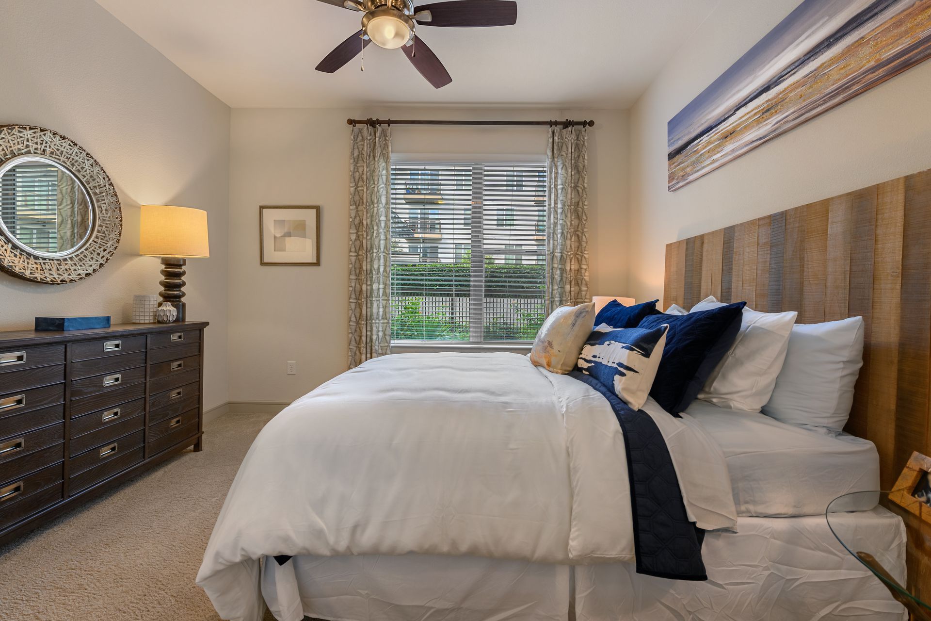A modern bedroom with a bed featuring a wood headboard, a white comforter, and dark blue pillows next to a dark dresser.