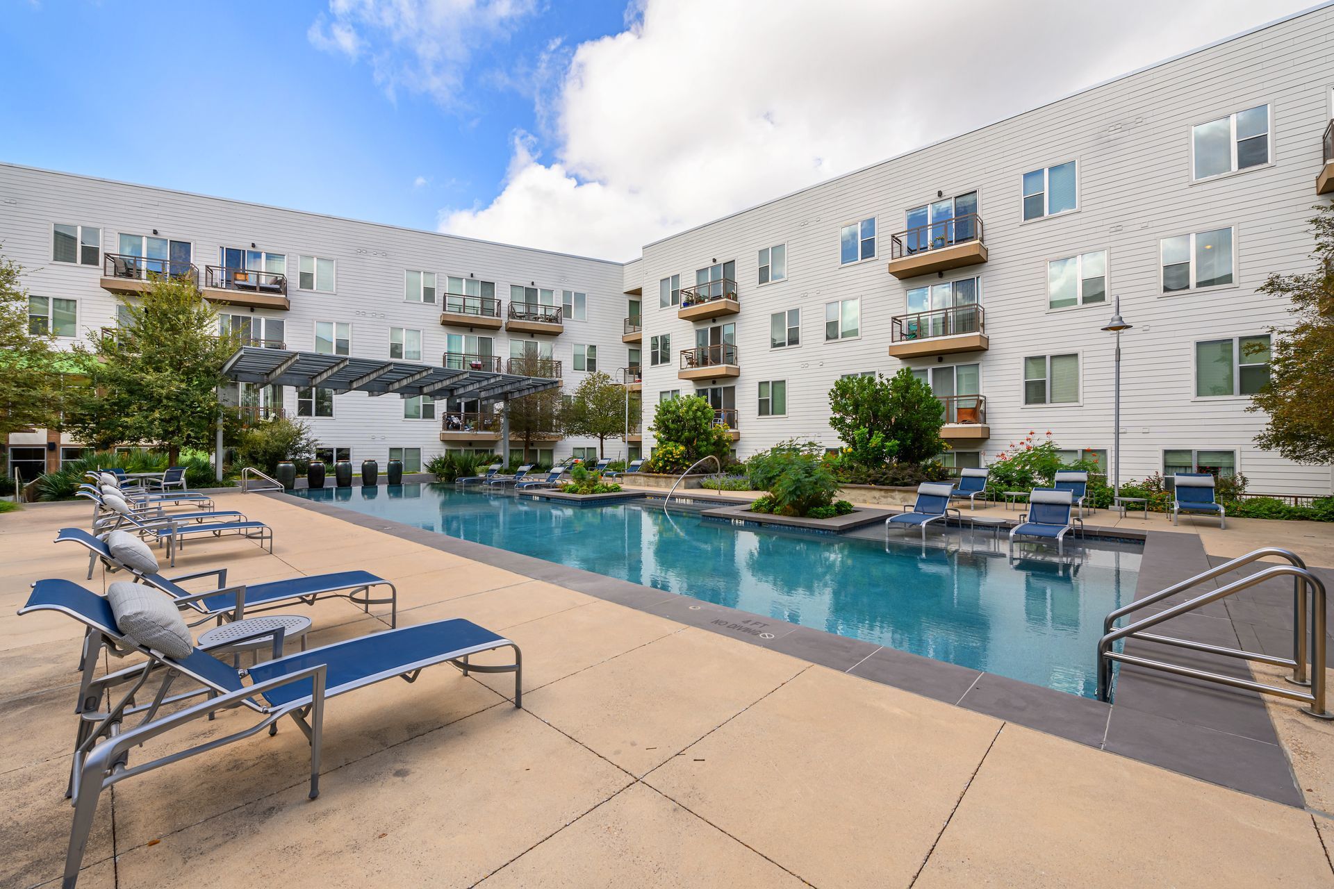 Outdoor swimming pool at an apartment complex, featuring lounge chairs on a concrete deck under a blue sky with clouds.