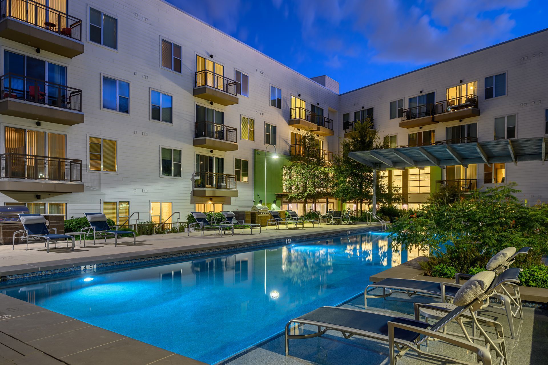 An illuminated apartment building pool area at dusk with lounge chairs on the deck and a small green space.